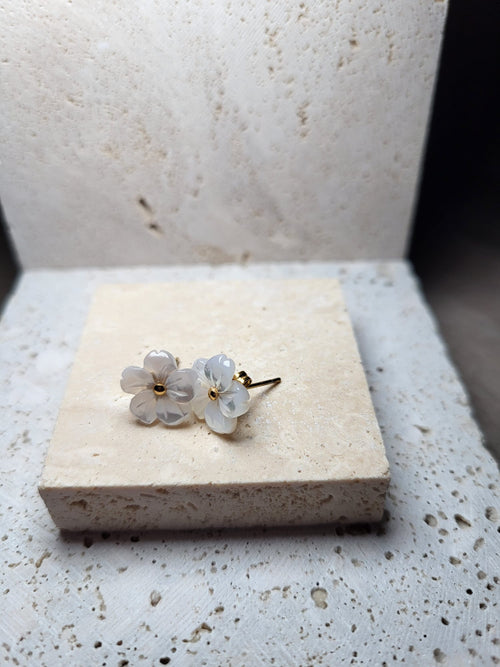 Twee delicate, parelmoeren bloem oorknopjes, gemaakt van stainless steel, liggen op een beige ondergrond.