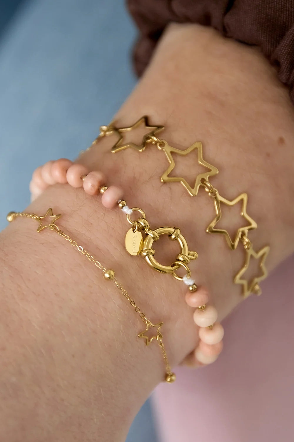 Arm met drie armbanden: gouden ster armband, Clarisa armband met roze koraal natuursteen kralen, en minimalistische gouden ster armband.