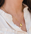 Gouden geboortesteen ketting met rondje verjaardagsbloem hanger en paarse steen als sieraad aan hals van vrouw in witte blouse