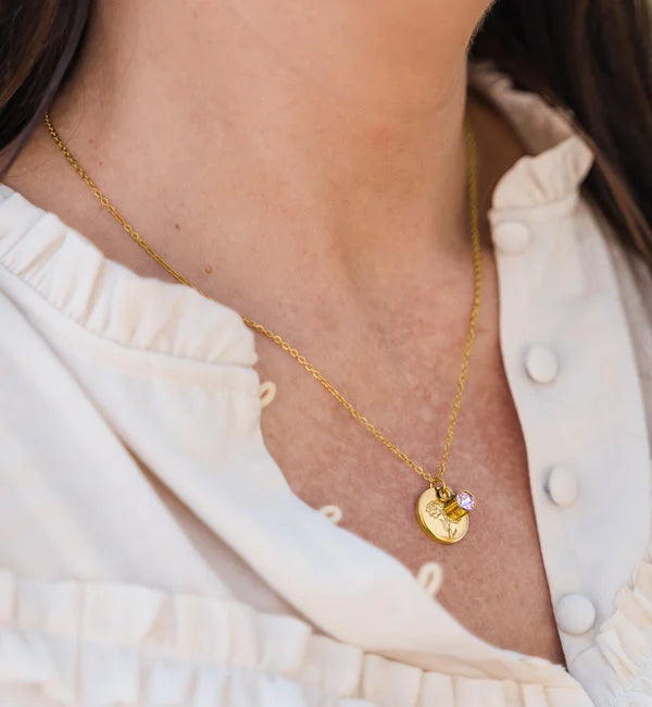 Gouden geboortesteen ketting met rondje verjaardagsbloem hanger en paarse steen als sieraad aan hals van vrouw in witte blouse