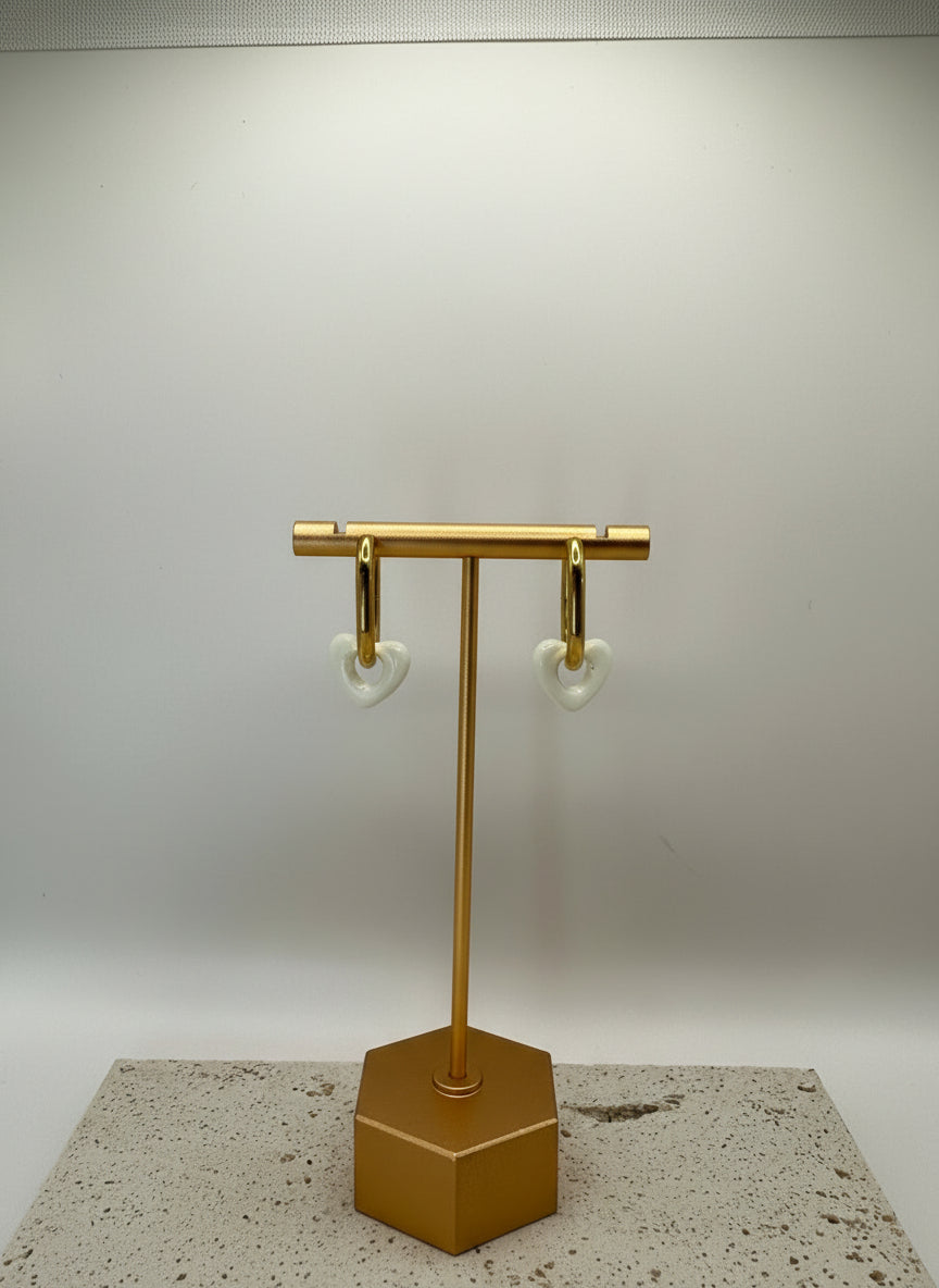 Gouden oorringen met witte hartjes van stainless steel, hangend aan een gouden display op een lichte ondergrond.