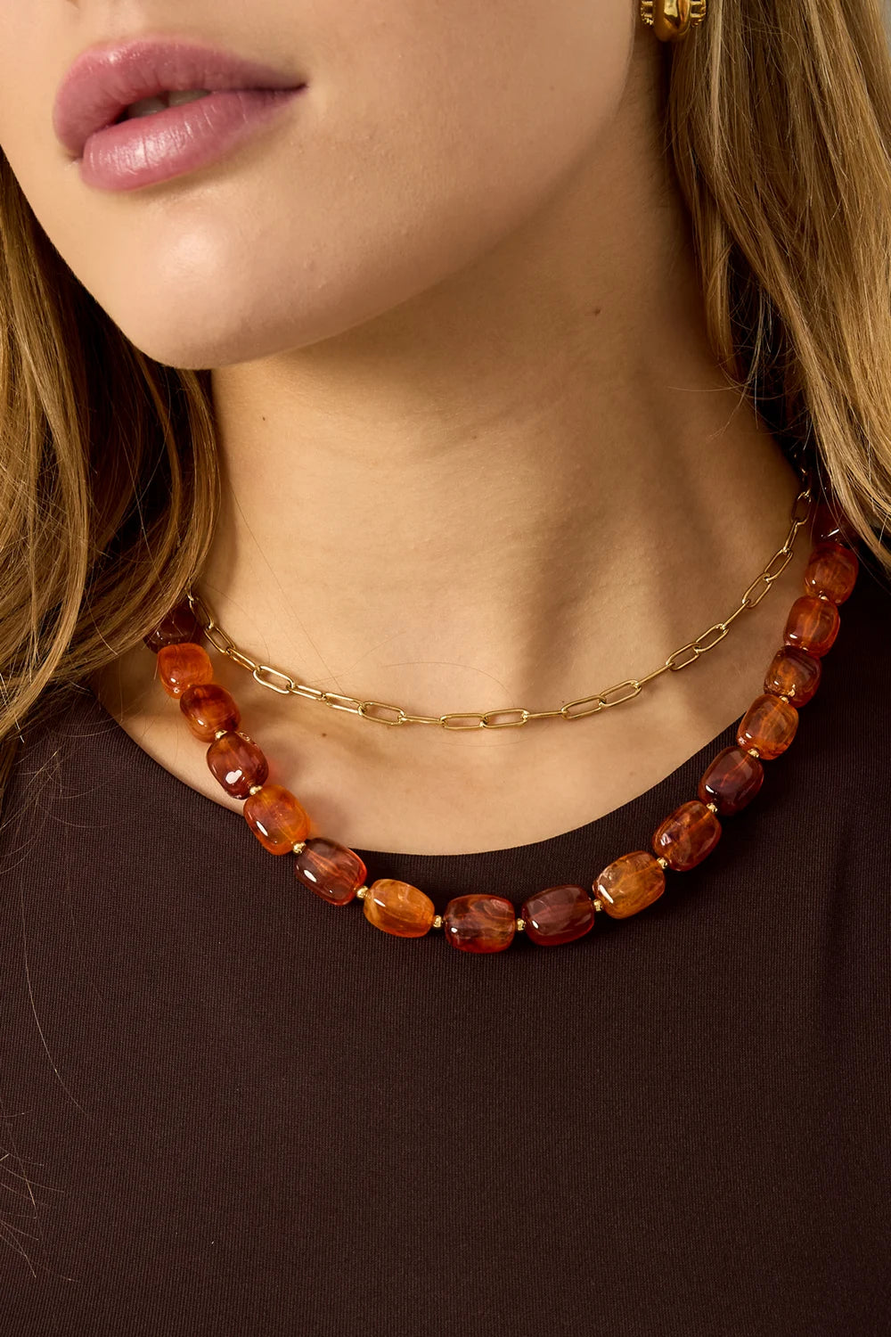Vrouw draagt dubbele ketting: goudkleurige schakelketting en Mara Ruby Red ketting met glanzende kunsthars kralen.