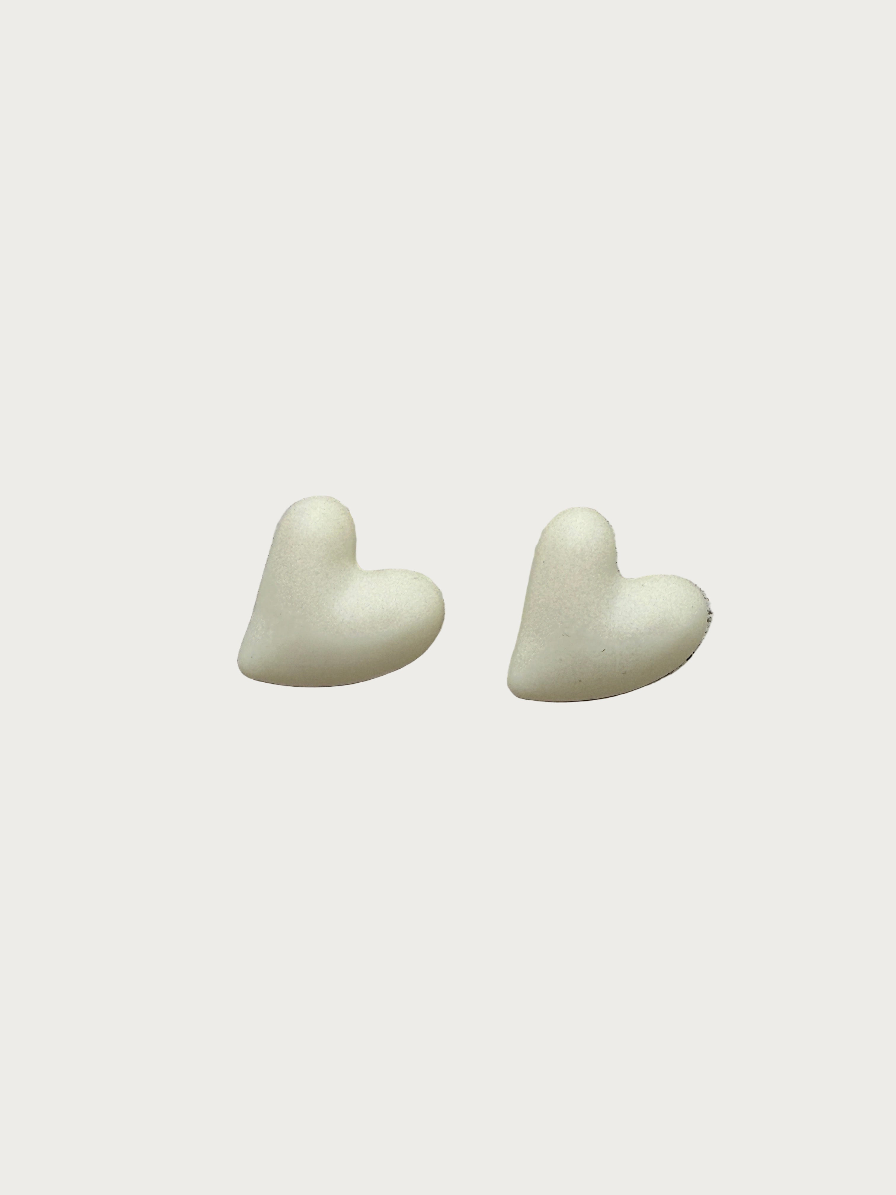 Twee lichtbeige hartjesoorbellen van polymeerhars, zachte studs met een matte textuur, perfect voor gevoelige oren.