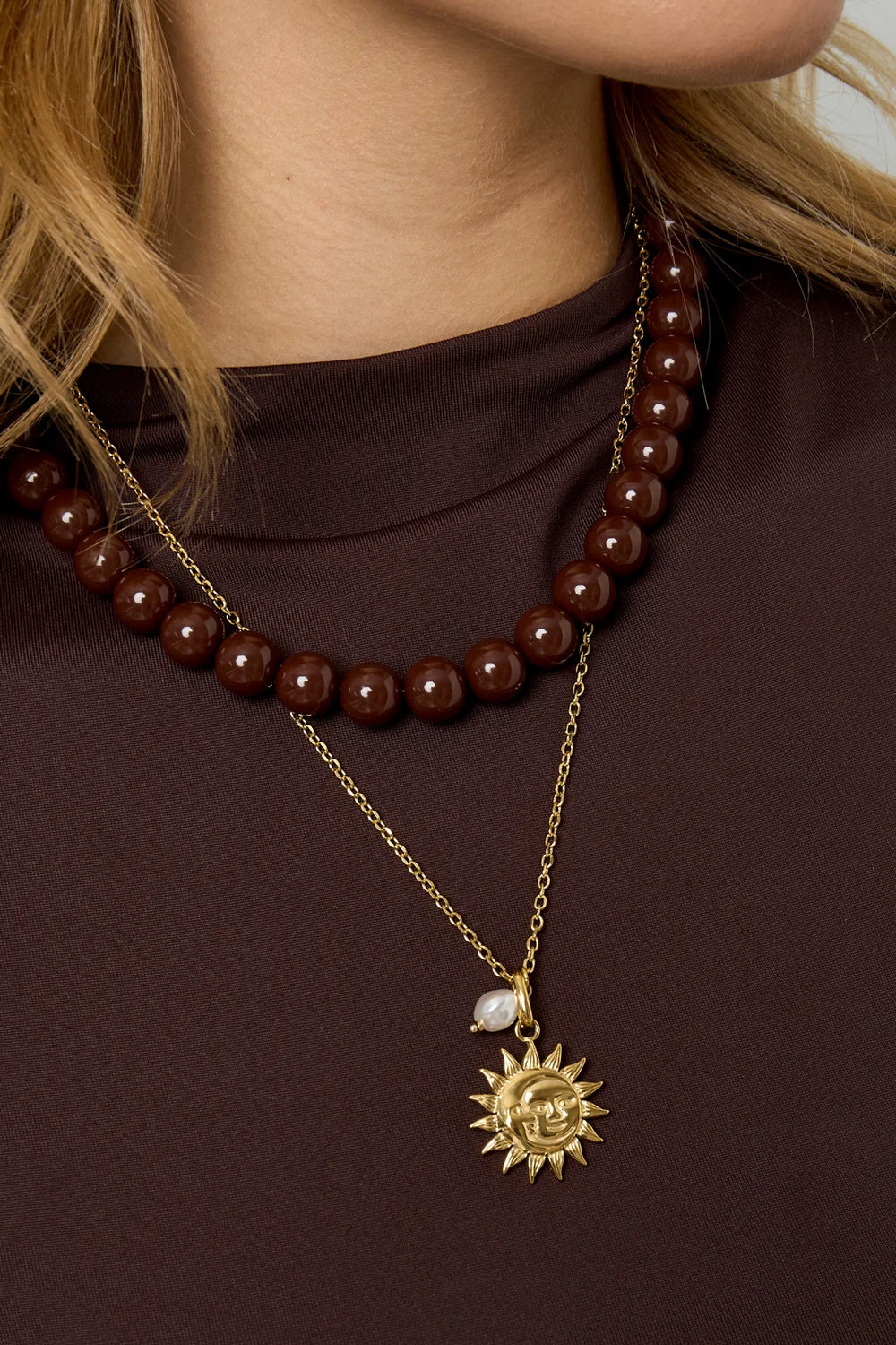 Gouden RVS ketting met zonne-hanger en parel, gecombineerd met bruine kralenketting op een model.