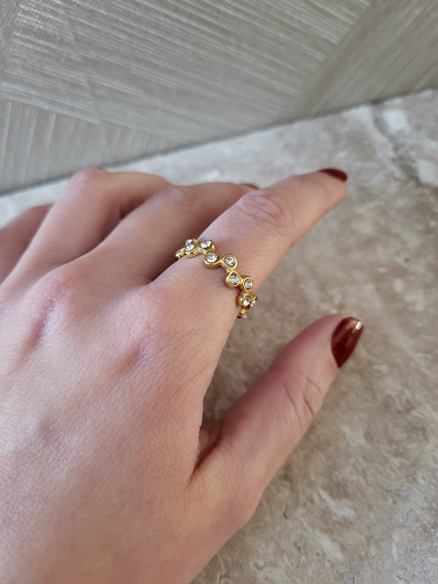 Gouden stainless steel ring met golfjes en ingelegde steentjes om een vinger, de hand heeft donkerrode nagellak