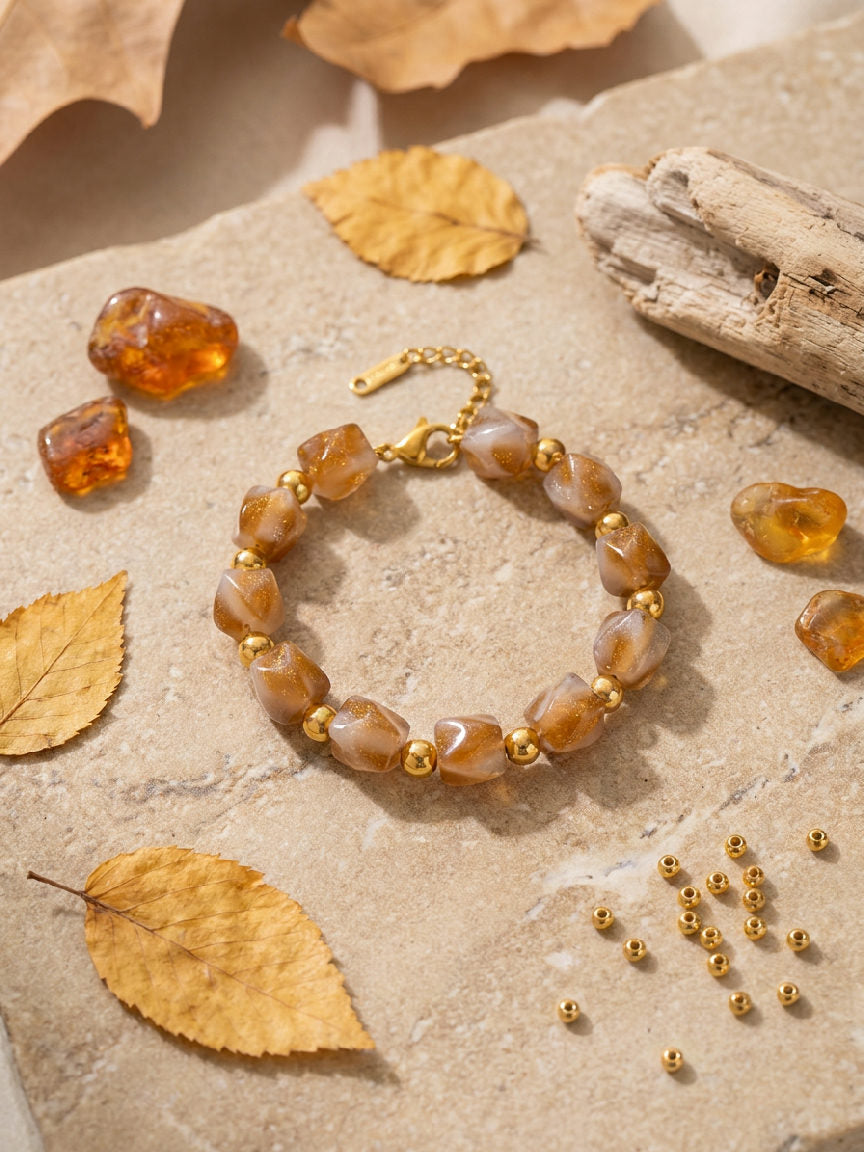 Amber kralenarmband met onregelmatige hars kralen en gouden details op een beige ondergrond met herfstbladeren en ruwe amberstenen.