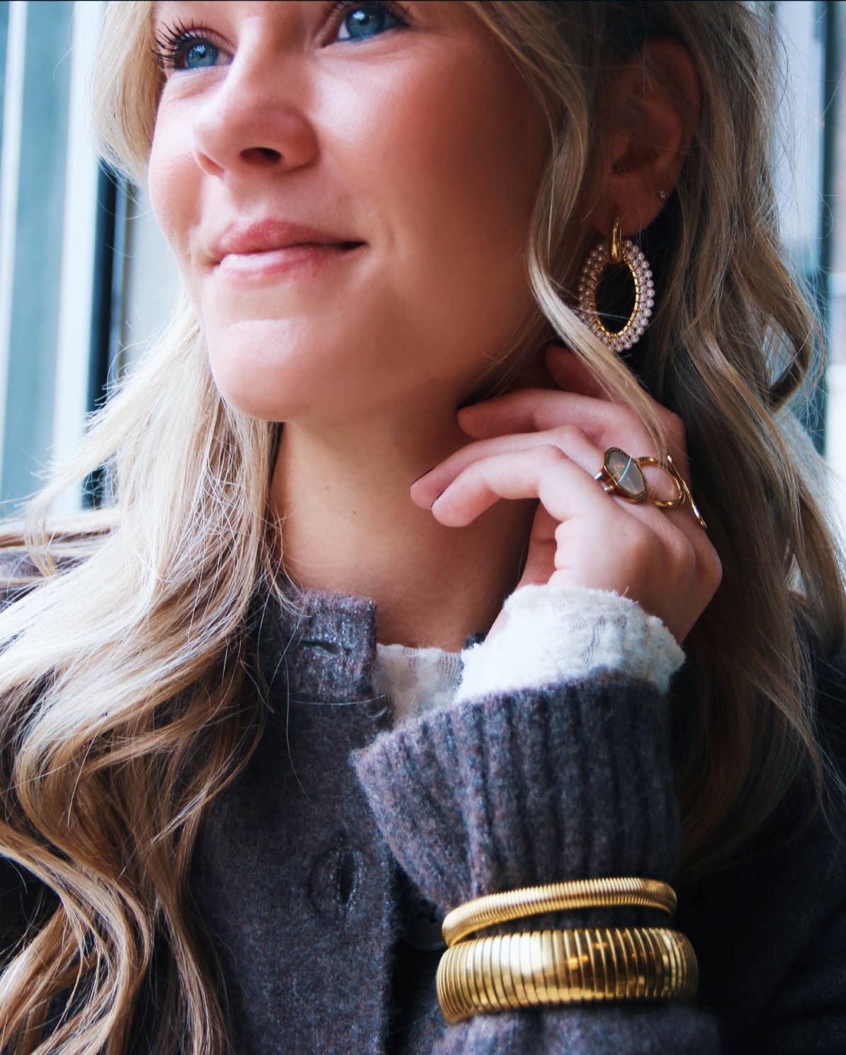 Vrouw met blonde haren draagt stainless steel Pearl Loop oorbellen met parels en gouden armbanden, elegant design.