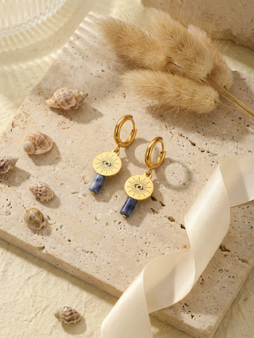 Gouden Luna oorbellen van stainless steel met een oogje en blauwe sodaliet steen aan hangers op een natuurstenen achtergrond.