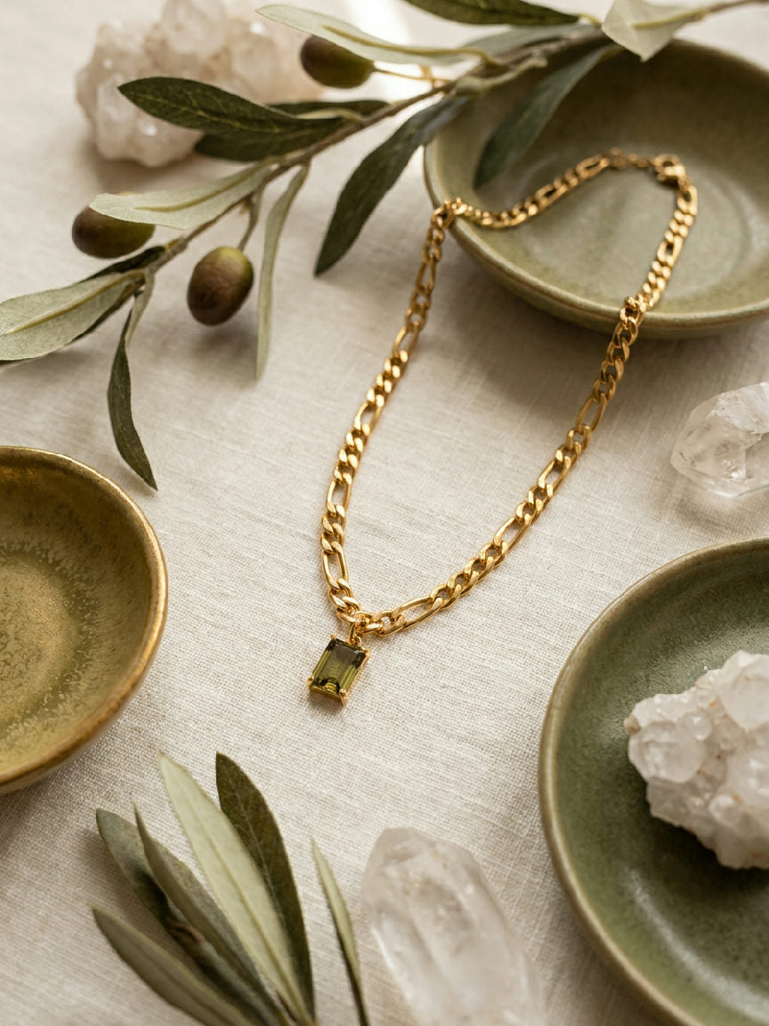 Gouden figaro ketting met rechthoekige groene steen hanger, gemaakt van stainless steel, op lichte ondergrond met olijftakken